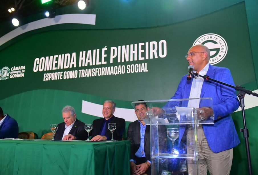 Homem de pé em tribuna e outros na bancada ao lado.