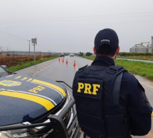 Policial da PRF ao lado de viatura em rodovia.