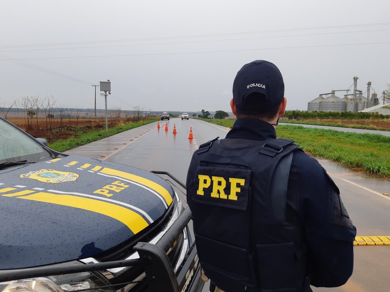 Policial da PRF ao lado de viatura em rodovia.